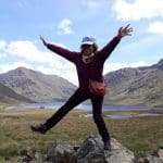 Person joyfully posing on rocks with arms outstretched in a green mountainous landscape. The green hills and blue lake behind the person are the perfect image of the rugged Irish countryside that Overland’s small-group tours of Ireland pass through.
