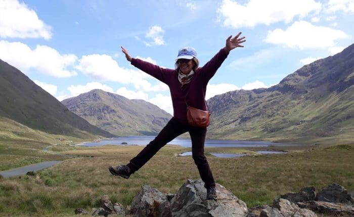 Person joyfully posing on rocks with arms outstretched in a green mountainous landscape. The green hills and blue lake behind the person are the perfect image of the rugged Irish countryside that Overland’s small-group tours of Ireland pass through.
