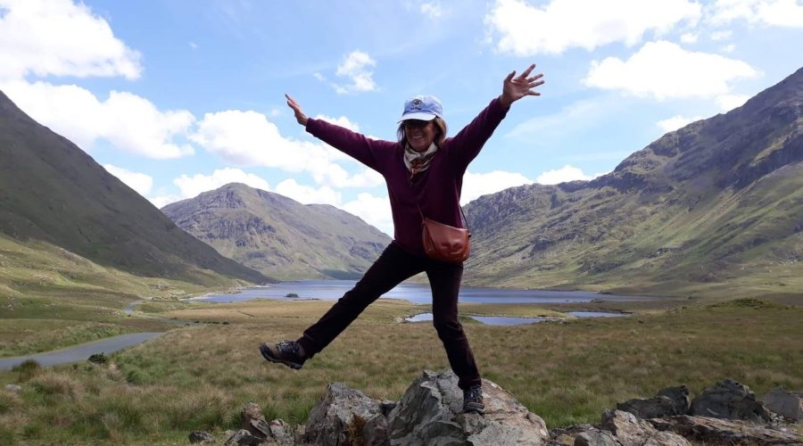 Person joyfully posing on rocks with arms outstretched in a mountainous landscape, with a lake and green hills in the background. Sky is partly cloudy and sunny, capturing the breathtaking beauty of a Northern Ireland tour.