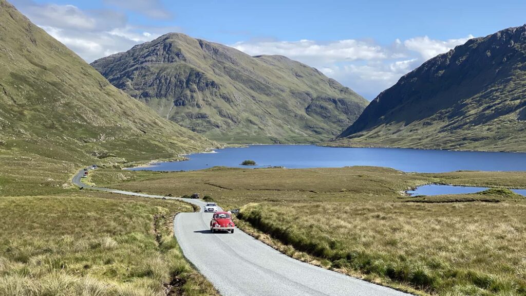 red car driving in road between green mountains and a lake