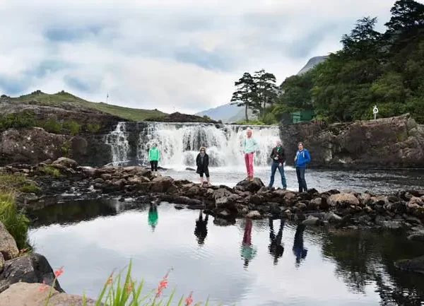 A group of tourists stands in front of cascading waterfalls with lush greenery, under a cloudy sky, reflected in a calm pool of water.