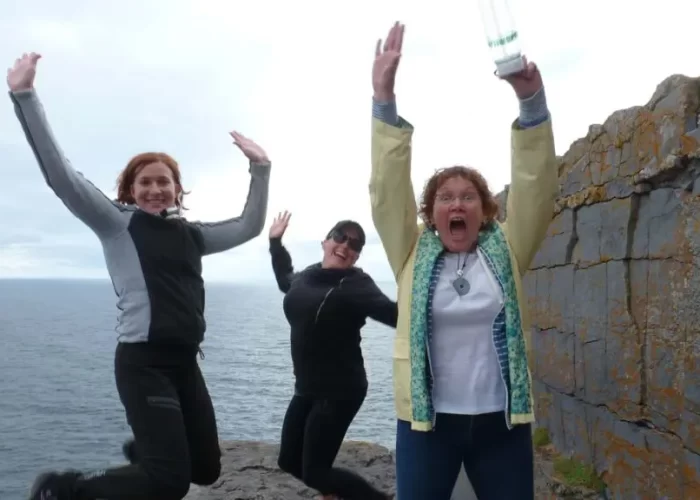 Three people are captured mid-jump near a rocky cliff with the ocean in the background. Two wear jackets and the third has a light-colored sweater. All appear to be excited and smiling.