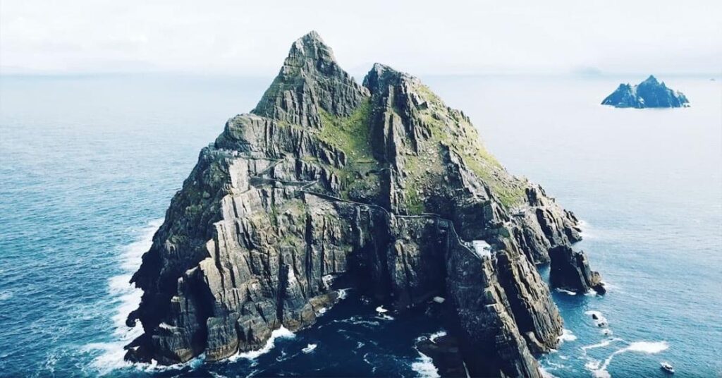 Aerial view of a rugged, steep island with sharp cliffs surrounded by the ocean, under a cloudy sky.