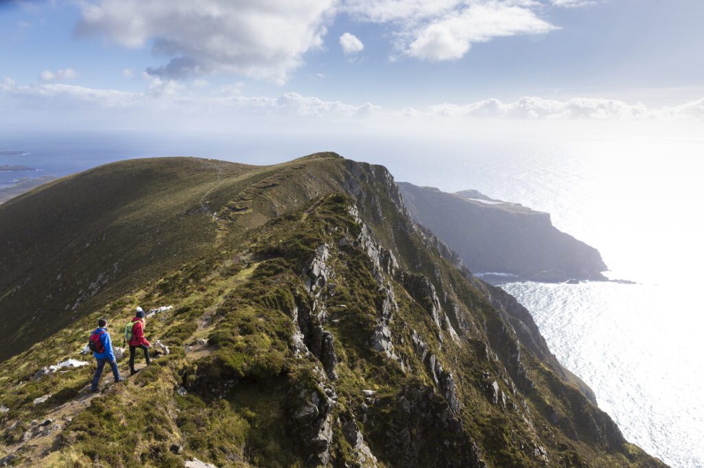 Two hikers walking on a mountain ridge overlooking the sea on a sunny day.