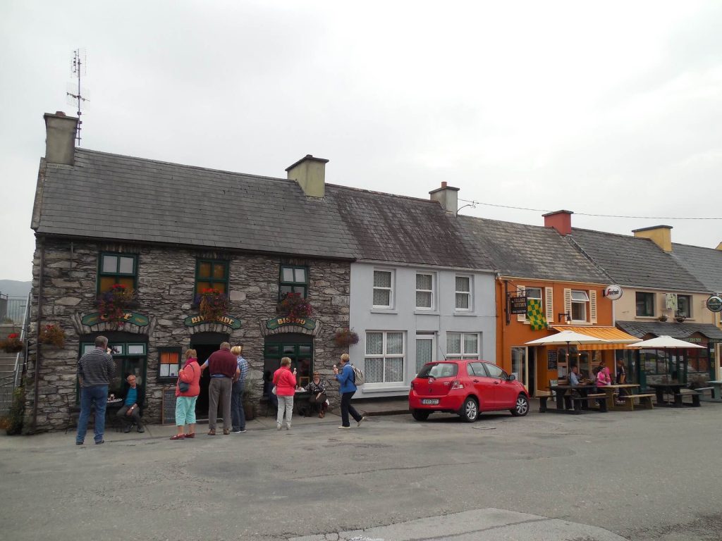 People standing outside a gray stone building with colorful adjoining shops and a red car parked in front.