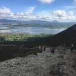 Hikers descending a rocky mountain trail with expansive views of a flat valley and distant mountains under a cloudy sky.