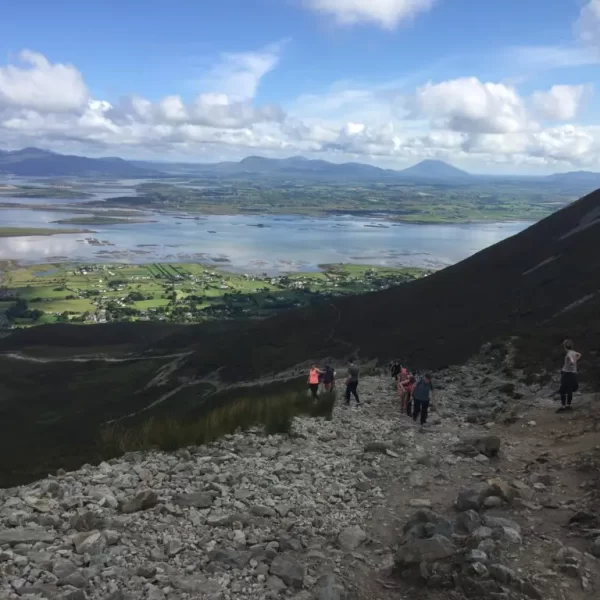 Hikers descending a rocky mountain trail with expansive views of a flat valley and distant mountains under a cloudy sky.