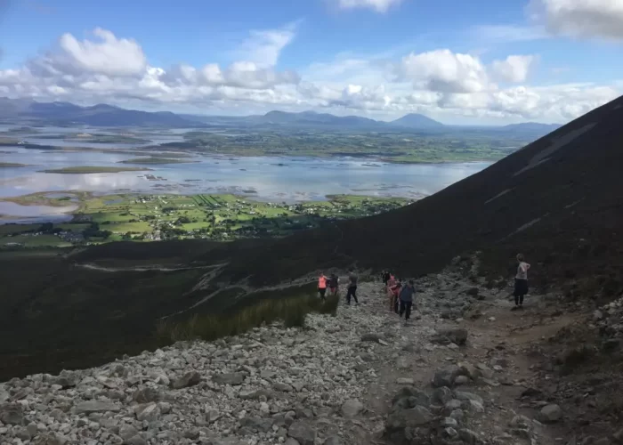 Hikers descending a rocky mountain trail with expansive views of a flat valley and distant mountains under a cloudy sky.