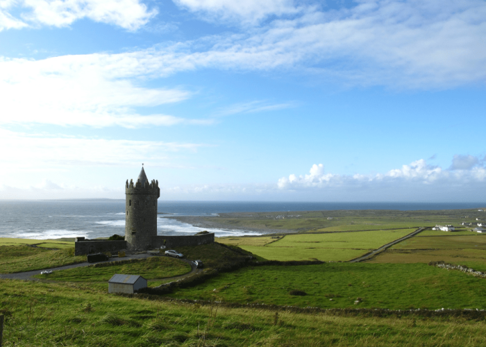 An ancient stone tower stands on a green hill overlooking a coastal landscape with the sea in the distance under a partly cloudy sky.
