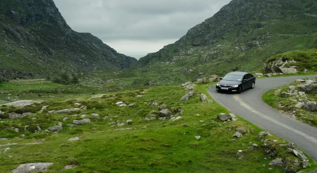 Car driving on a winding road through a rocky, green mountain pass under an overcast sky.