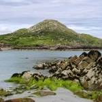 A serene coastal landscape featuring a grassy hill, rocky shoreline, and clear blue water under a cloudy sky.
