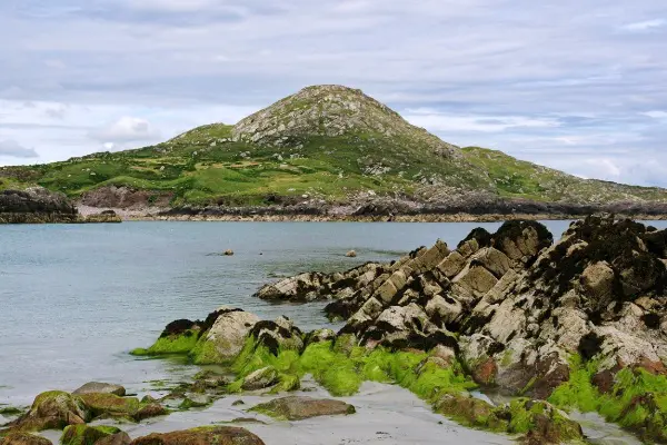 A serene coastal landscape featuring a grassy hill, rocky shoreline, and clear blue water under a cloudy sky.