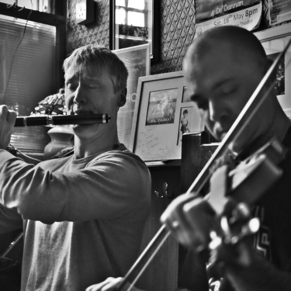 Two musicians performing in a dimly lit pub, one playing the flute and the other the violin, with posters in the background.