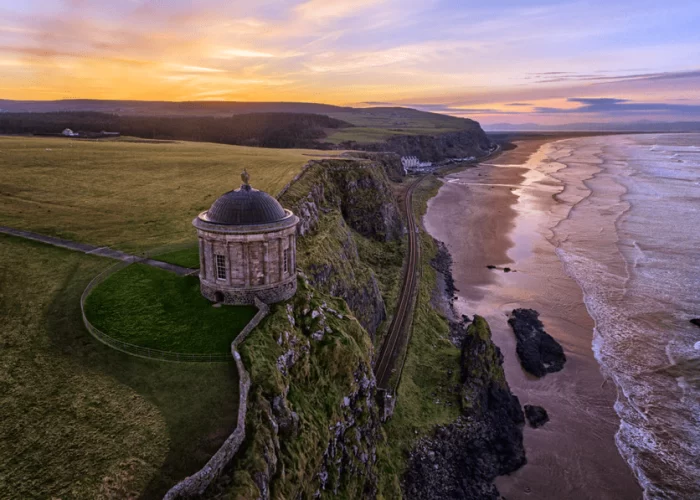 Aerial view of mussenden temple on a cliff overlooking a beach at sunset, with waves gently lapping the shore and expansive grassy fields surrounding the structure.