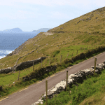 A narrow road winding along a lush green hillside beside a rocky coastline with the ocean in the background.
