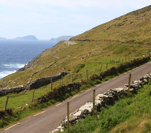 A narrow road winding along a lush green hillside beside a rocky coastline with the ocean in the background.