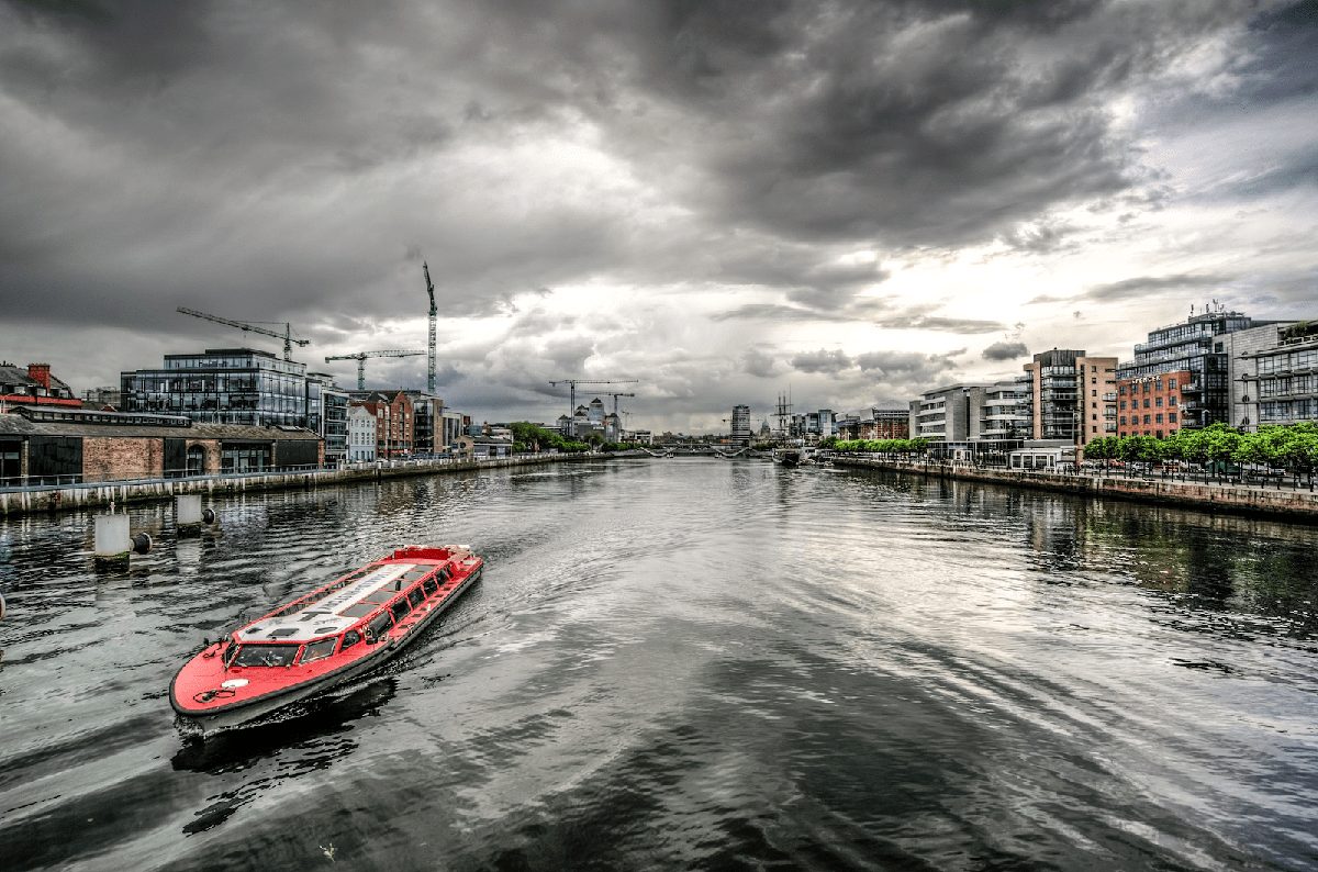 A red tour boat sails along a river flanked by modern buildings under a cloudy sky.