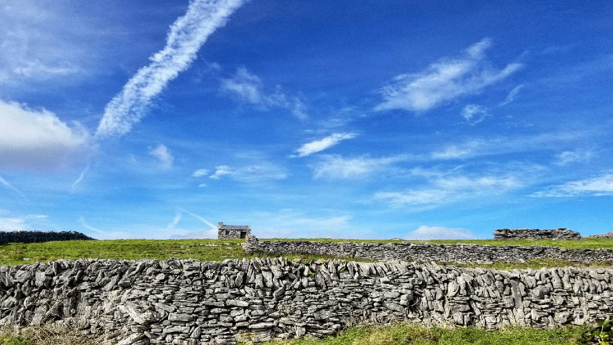 Stone walls and a solitary stone house in a lush green field under a blue sky with wispy clouds.