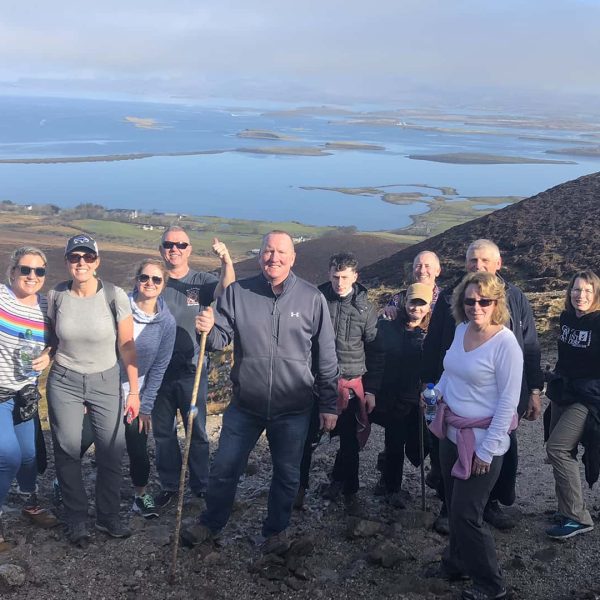 A group of people, some with hiking gear, stand on a rocky hillside with a scenic view of a body of water and small islands in the background, enjoying one of our tailor-made tours.