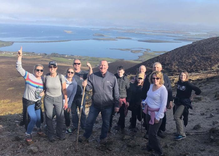A group of people, one with a hiking stick, stand on a rocky hillside with a scenic view behind them. They are on high ground, on the side of a mountain, and the lower ground stretches out below them before giving way to the sea and various islands. This kind of beautiful Irish countryside is at the heart of Overland’s Ireland tours.
