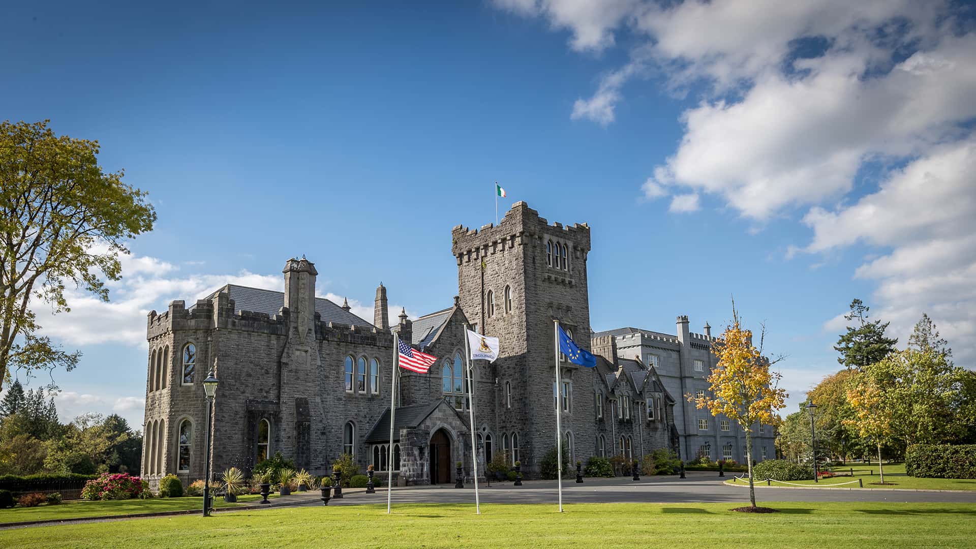 A large stone castle with turrets and flags in front, set against a blue sky with a few clouds and surrounded by grass and trees.