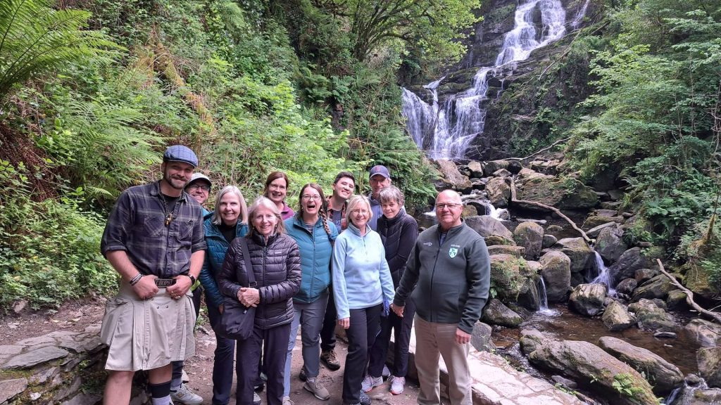 A group of people stand on a trail in front of a waterfall, surrounded by lush greenery, during one of Ireland's private guided tours.