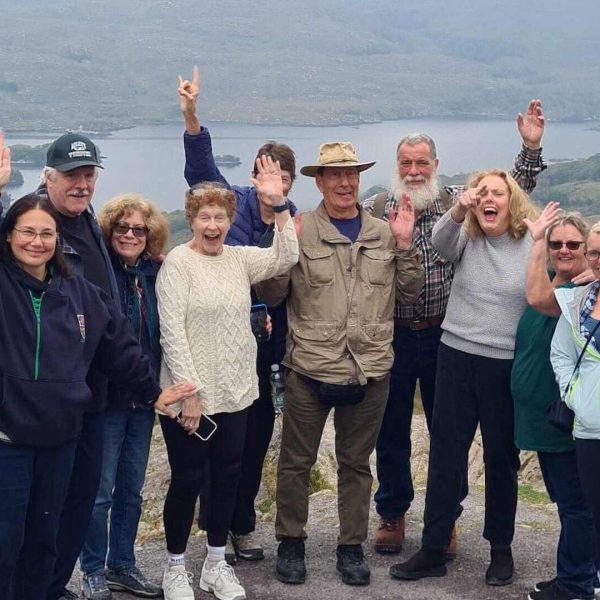 A group of ten people stands outdoors on a trail with a scenic backdrop of mountains and a lake, all smiling and waving at the camera during their Private Ireland Tour.