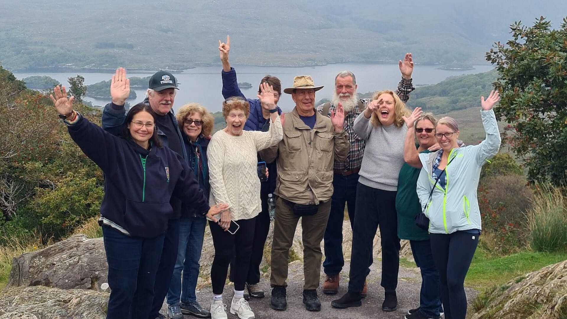 A group of ten people stands outdoors on a trail with a scenic backdrop of mountains and a lake, all smiling and waving at the camera during their Private Ireland Tour.