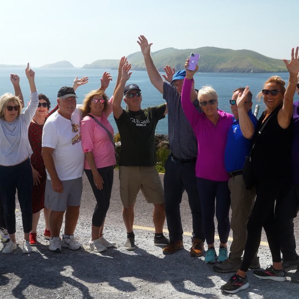 A small group of people stands in front of a coastal landscape with hands raised, smiling at the camera, embracing the spirit of adventure on one of their tours of Ireland.