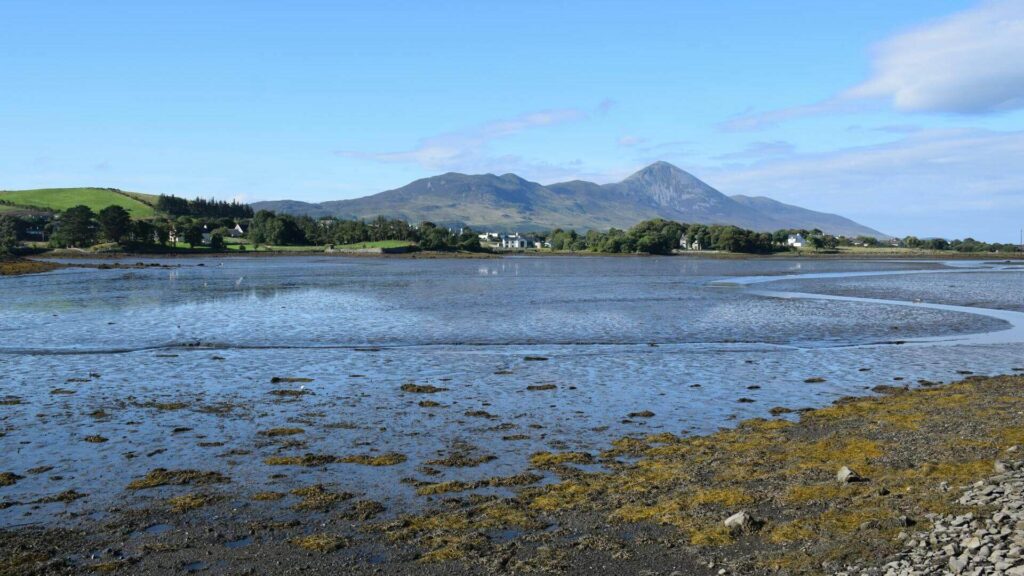 Scenic view of a shallow tidal waterway with rocky foreground, green hills, and distant mountains under a clear blue sky.