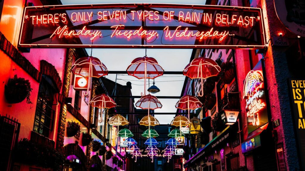 Colorful neon lights in an alley display umbrellas and a sign that reads, "There's only seven types of rain in Belfast: Monday, Tuesday, Wednesday...