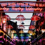 Colorful neon lights in an alley display umbrellas and a sign that reads, "There's only seven types of rain in Belfast: Monday, Tuesday, Wednesday...