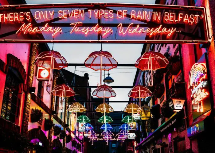 Colorful neon lights in an alley display umbrellas and a sign that reads, "There's only seven types of rain in Belfast: Monday, Tuesday, Wednesday...