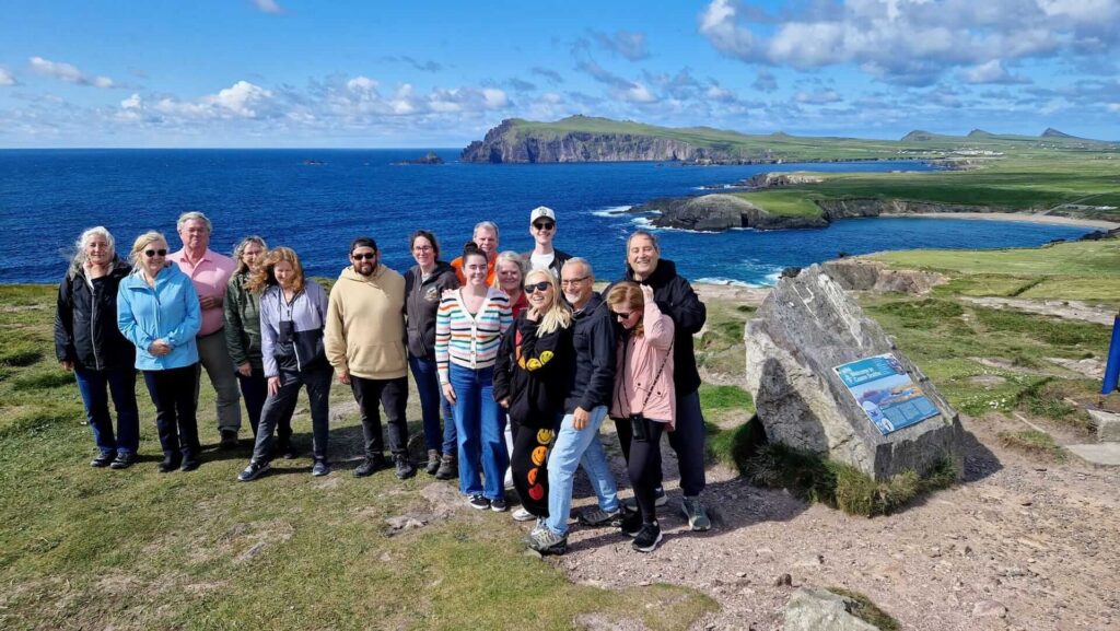 A group of people stands on a grassy cliff, basking in the ocean views and distant coastline—an idyllic scene often enjoyed on Ireland Tours. A stone plaque is visible on the right, marking this spot as a cherished highlight of small group tours.