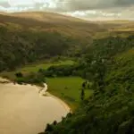 Aerial view of a lake bordered by forested hills and grassy fields under a cloudy sky.