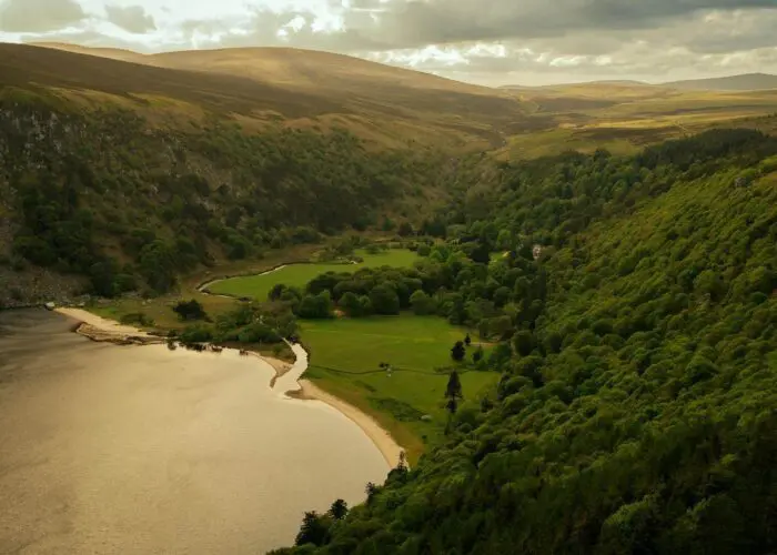 Aerial view of a lake bordered by forested hills and grassy fields under a cloudy sky.