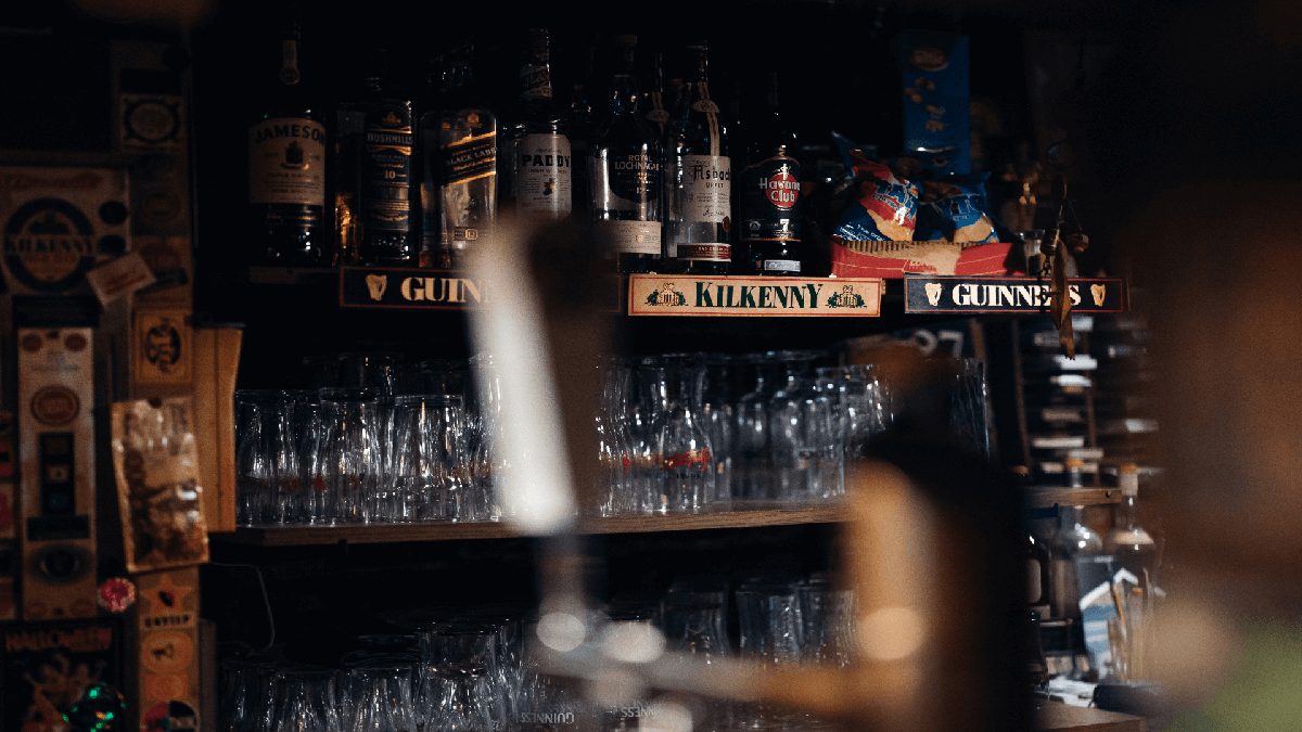 Bar shelf with various liquor bottles and upside-down glasses. Kilkenny and Guinness branding visible.