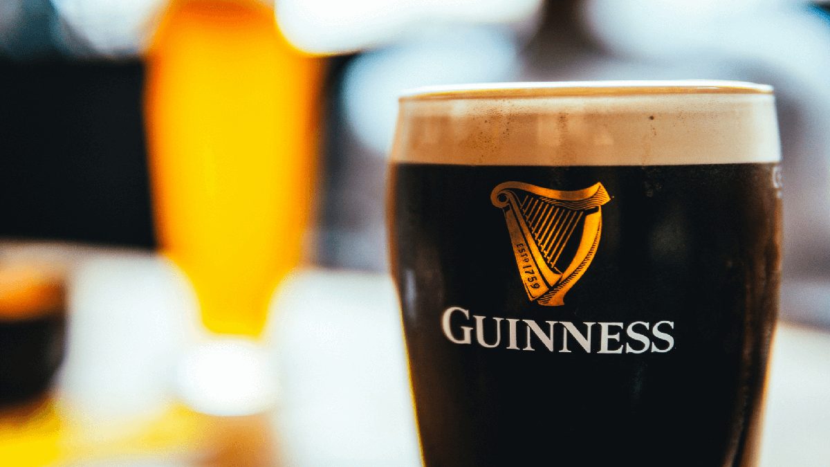 Close-up of a pint of Guinness with a foamy head, logo visible on the glass. A background of blurred warm light and a second illuminated drink is partially visible.