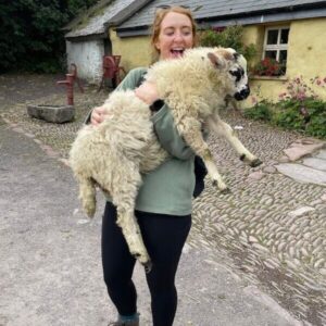 Person holding a sheep outside a rustic building with cobblestone path.